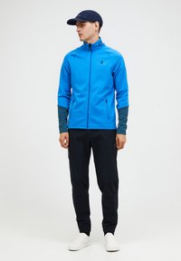 Giacca blu con zip e maniche grigie scure, abbinata a pantaloni neri e sneakers bianche. Tessuto liscio; design funzionale con tasche laterali.