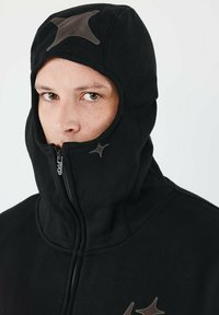 Sort hættetrøje i fleece med højt krave og lynlås. Har stjerneformede brune accenter. Blødt materiale, designet til varme.