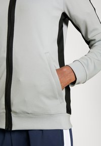Lichtgrijze zip-up jack met zwarte zijpanelen, ribgebreide manchetten en een voorkeep. Glad textuur met zichtbare stikseldetails.