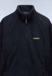 Pullover in pile di colore blu navy con colletto alto, chiusura a mezza zip e tasche laterali. Presenta una piccola toppa con logo sul petto. Tessuto morbido.