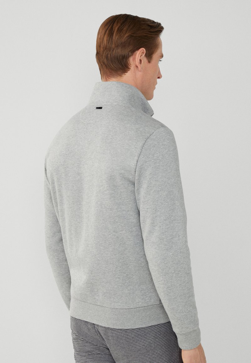 Mann mit braunen Haaren, der einen hellgrauen langärmeligen Pullover mit Stehkragen und gemusterten Hosen trägt, mit dem Rücken zur Kamera.