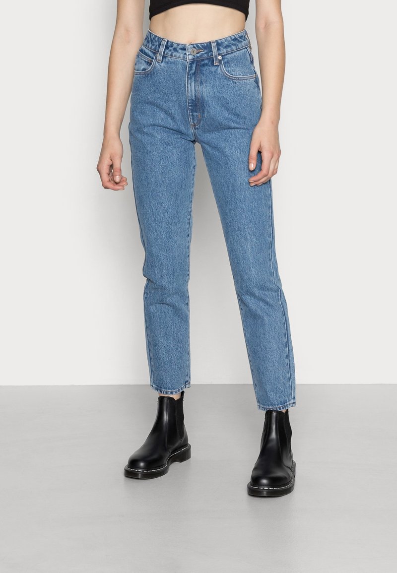 Femme portant un jean bleu taille haute et des bottines noires, debout devant un fond clair uni.