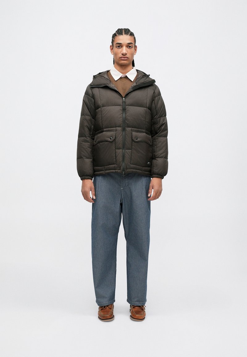 Brun polstret jakke med hette, glidelås og to frontlommer. Brukt over en brun genser og lyseblå, løstsittende jeans.