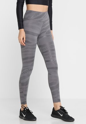 Leggings de sport gris avec un motif ondulé et un design texturé, taille haute, fabriqués à partir d'un matériau extensible, associés à des baskets noires.