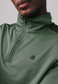 Sudadera verde con cremallera y cuello alzado, con tejido texturizado, suturas sutiles y un pequeño logo en el pecho.