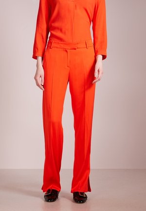 Rode op maat gemaakte broek met een rechte pijp, gemaakt van gladde stof. Kenmerkt zich door plooien aan de voorkant en een riem tailleband. Draag met zwarte schoenen.