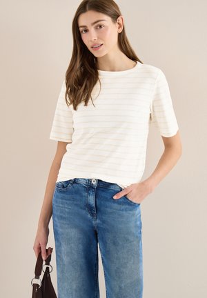 Giovane donna che indossa una maglietta bianca a righe con maniche corte e jeans blu, tenendo in mano una borsa marrone con dettaglio ad anello.