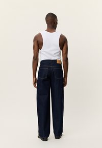 Σκούρα μπλε τζιν παντελόνια με φαρδιά μπατζάκια από τζιν ύφασμα, με δερμάτινο ταμπελάκι ταμπά στην πίσω μέση. Φορεμένα με λευκή αμάνικη μπλούζα.