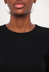 Haut noir à col rond porté avec de grandes boucles d'oreilles en argent brillantes. Texture du tissu lisse, mettant en valeur un gros plan du décolleté et des bijoux.