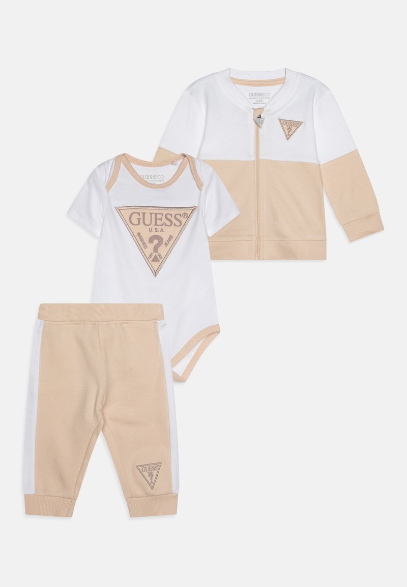 Třídílný kojenecký outfit zahrnuje béžovou a bílou zipovou bundu, bílý body s béžovým logem a béžové kalhoty s bílými akcenty.
