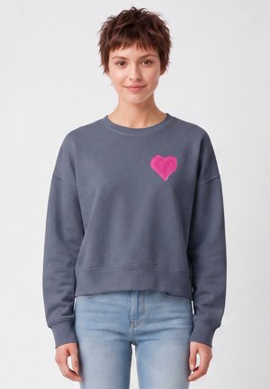 Jeune personne aux cheveux courts portant un sweat-shirt gris avec un cœur rose sur la poitrine et un jean bleu clair, souriant légèrement.