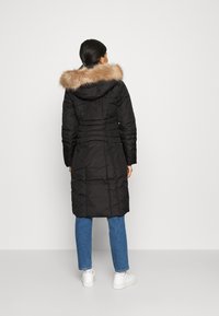 Svart isolerad jacka med huva av syntetpäls, strukturerad quiltning och lång längd. Kombineras med blå jeans och vita sneakers.