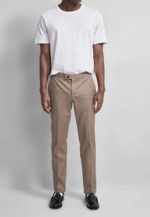 Uomo che indossa una maglietta bianca a maniche corte semplice, pantaloni beige slim fit e mocassini neri in pelle, in piedi contro un muro grigio.