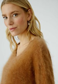 Brauner Mohair-Mix-Pullover mit einer weichen, kuscheligen Textur und V-Ausschnitt. Das Model trägt Perlenohrringe und präsentiert einen natürlichen Make-up-Look.