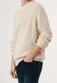 Pull en tricot couleur crème avec encolure et poignets côtelés, doté d'une coupe décontractée et d'un tissu texturé, assorti à un jean en denim foncé.