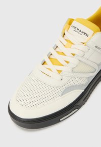 Witte sneaker met gele accenten, geperforeerde bovenkant, grijze details en zwarte zool. Beschikt over een textuurstof tong en veters.