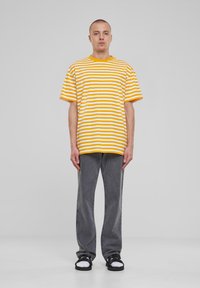 Mann steht mit einem gelb-weiß gestreiften T-Shirt, grauen Jeans, weißen Socken und schwarzen Sandalen vor einem schlichten grauen Hintergrund.