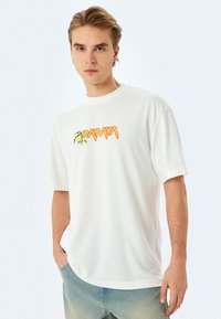 Hvid kortærmet T-shirt lavet af blødt stof, med et farverigt "PAPAYA" logo med grønne og orange detaljer, afslappet pasform.