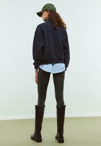 Námornícka oversized mikina preložená cez svetlomodrú prúžkovanú košeľu, doplnená tmavozelenými legínami a čiernymi čižmami po kolená. Zelená čiapka.