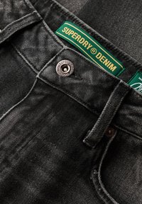 Jeans de mezclilla negra con un acabado texturizado, que presentan una etiqueta verde con "SUPERDRY DENIM", un botón metálico y costuras resistentes.