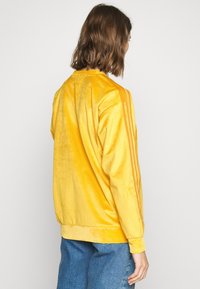 Gul manchesterjacka med ribbad struktur, tre vertikala ränder på ärmarna och en avslappnad passform. Bakifrån syns detaljer på kragen och mudden.