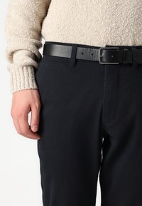 Crne teksturirane hlače slim fit kroja, uparene s glatkim crnim kožnim remenom s kvadratnom kopčom, nosi se s bež pletenim puloverom.