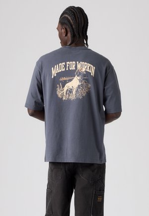 Uomo con capelli intrecciati che indossa una maglietta grigio scuro con un cane e il testo "Made for Workin" sulla schiena, abbinata a pantaloni neri.