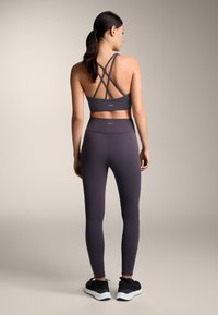 Kvinna stående med ryggen vänd, iklädd en mörklila sportbh med korsade band, matchande tights och svarta träningsskor på neutral bakgrund.