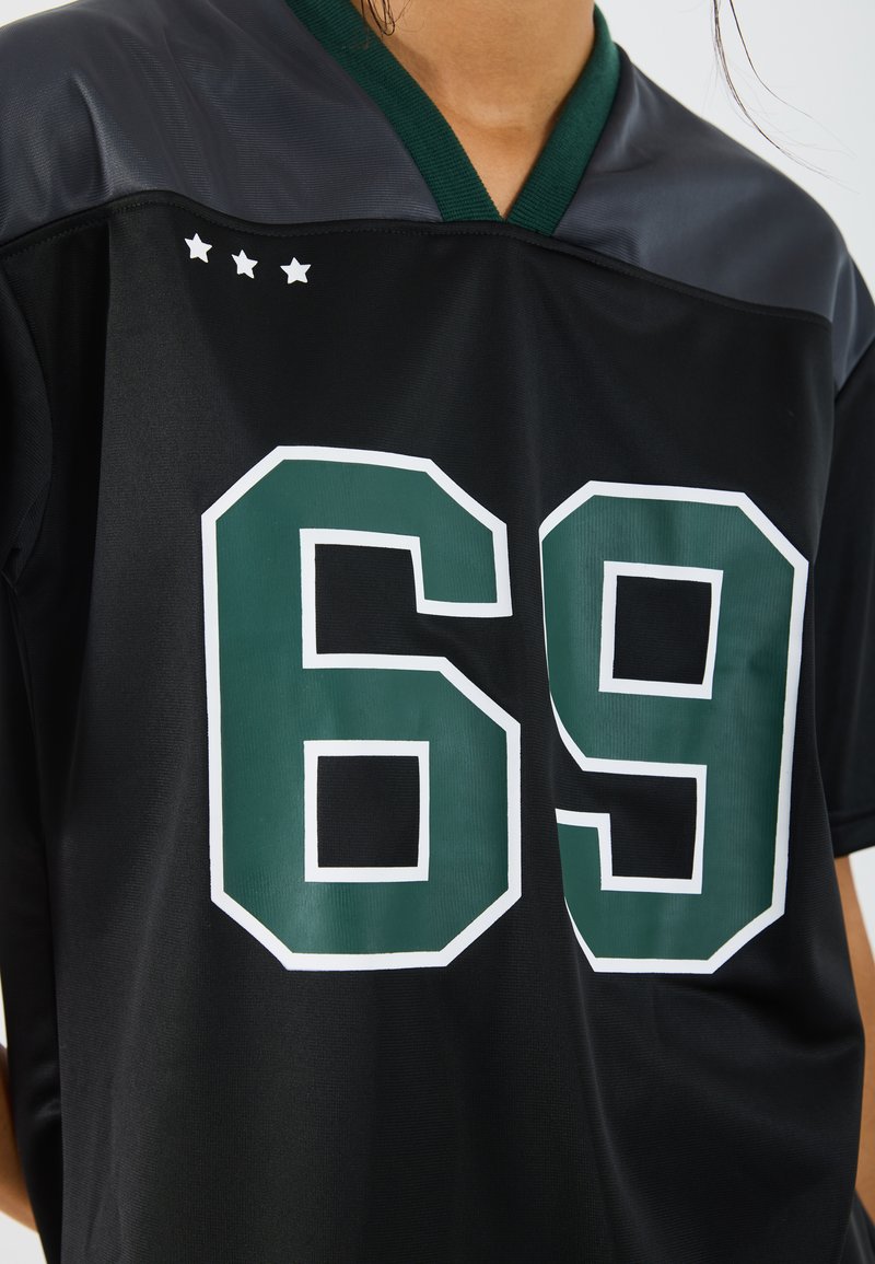 Camisa negra con el número verde "69" y tres estrellas blancas en el hombro izquierdo. Presenta un escote en forma de V y una textura deportiva.