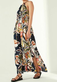 Robe maxi florale avec un col halter, présentant un fond marine et des imprimés botaniques colorés. Fente sur le côté et sandales à lanières noires.