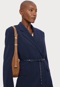 Ναυτικό σακάκι με δομημένα πέτα, διακοσμημένο με μια μαύρη δερμάτινη ζώνη. Καφέ σουέτ τσάντα με ρυθμιζόμενο λουράκι resting on the shoulder.