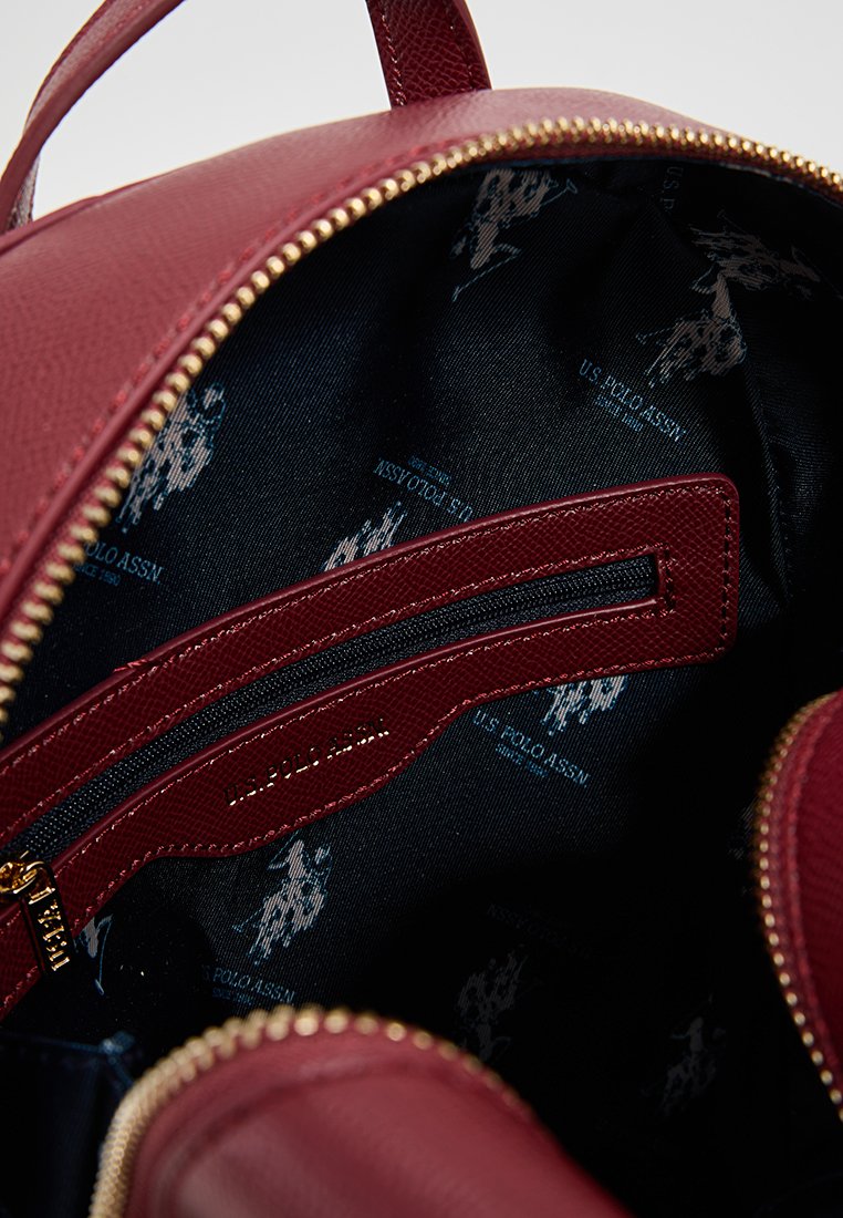 Interno di uno zaino in pelle borgogna con una tasca con zip, caratterizzato da un rivestimento stampato in blu navy con loghi ripetuti e una cerniera dorata.