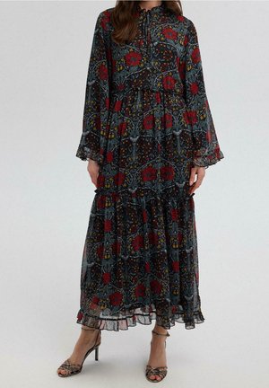 Frau trägt ein langes, dunkles Blumenkleid mit roten und gelben Blumen, langen Ärmeln, einem Rüschensaum und hochhackigen Sandalen mit Riemchen.