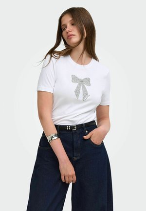 Kvinde iført hvid T-shirt med sølv sløjfe-design, mørke jeans med højtalje, sort bælte og sølv manchetarmbånd, poserer med den ene hånd i lommen.