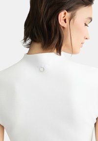 Top corto bianco a maniche corte con un collo alto e dettaglio con bottoni nella parte posteriore. Il tessuto appare testurizzato e morbido.