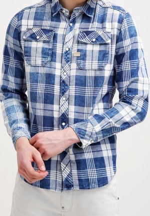 Homme portant une chemise à manches longues à carreaux bleus et blancs avec deux poches poitrine, ajustant le poignet, vêtu de pantalons blancs.