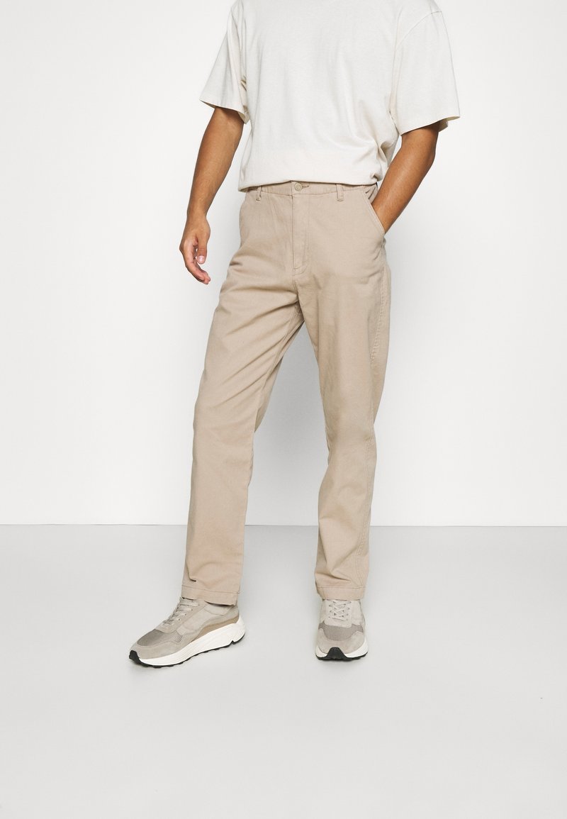 Personne portant un pantalon beige droit, un t-shirt blanc et des baskets beiges, debout avec une main dans la poche sur fond blanc.