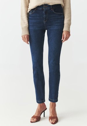 Kvinde iført mørkeblå slim-fit jeans, beige sweater og brune høje hælede sandaler med åben tå, stående foran en hvid baggrund.