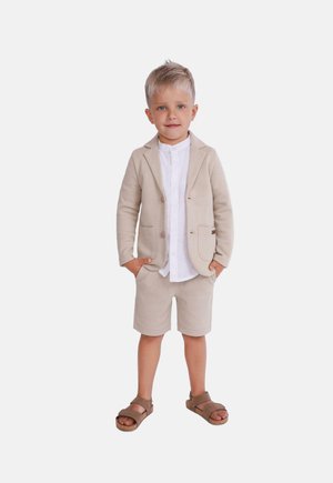 Bežové sakko na míru přes bílé košili s knoflíky, doplněné o bežové šortky a hnědé sandály. Hladká tkanina a strukturovaný design.