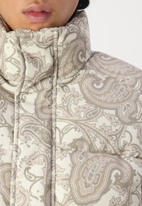 Puffer jacket with a high collar, featuring a beige paisley pattern on textured fabric. Stitching detail visible along seams and edges.