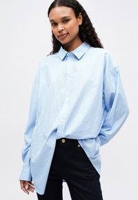 Lys blå stripete skjorte med krage, oversized passform, lange ermer og buet nederkant, brukt over mørke jeans.