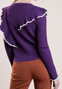 Pull violet côtelé avec accents à volants blancs. Manches longues ajustées et col rond. Tissu texturé qui ajoute de la profondeur.