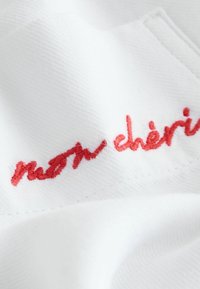 Belo blago z strukturirano tkanino z rdečim vezenim napisom "mon chéri." Šivi so natančni in rahlo izpostavljeni.