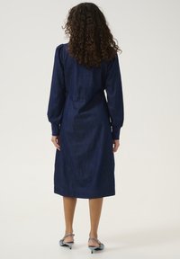 Jeansklänning i mörkblått med långa, puffiga ärmar och insvängd midja. Knä-length med rak skärning och minimal sömdetaljering.