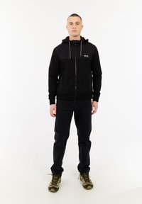 Man die met het gezicht naar voren staat, gekleed in een zwarte ritsvest, zwarte broek en groene en zwarte wandelschoenen op een witte achtergrond.