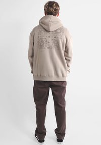 Haut à capuche beige avec un grand design graphique au dos, associé à un pantalon brun foncé et des baskets noires. Texture douce, coupe ample.