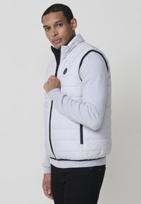 Weißer, gesteppter Puffer-Weste mit hohem Kragen und Reißverschluss. Enthält schwarze Akzente und ein strukturiertes graues Langarmshirt darunter.