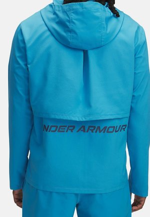 Person trägt eine hellblaue Under Armour Jacke mit Kapuze, sichtbares schwarzes Textlogo auf dem Rücken, steht mit dem Rücken zur Kamera.