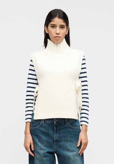 Λευκό ριμπαριστό αμάνικο τοπ με ψηλό γιακά, επάνω από μπλε και λευκό ριγέ μακρυμάνικο πουκάμισο, σε συνδυασμό με φαρδιά τζιν παντελόνια.