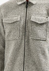 Grijze fleecetruffel met een ritssluiting aan de voorkant, twee borstzakken en een kraag. Het materiaal heeft een gestructureerde, zachte uitstraling.